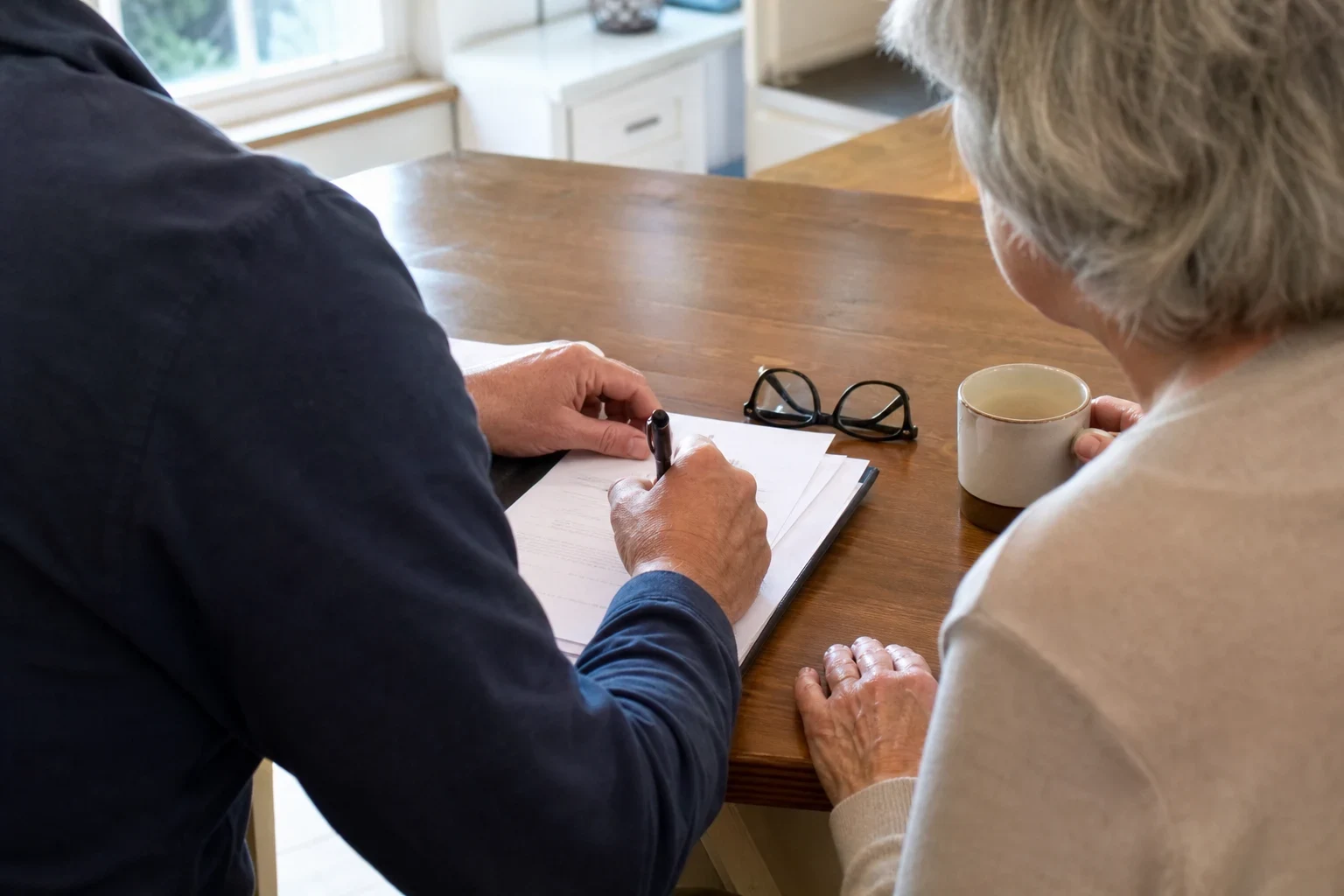 Over-the-shoulder view of an agent writing on a paper form at a wooden kitchen table beside a gray-haired woman holding a coffee mug, reading glasses on the table